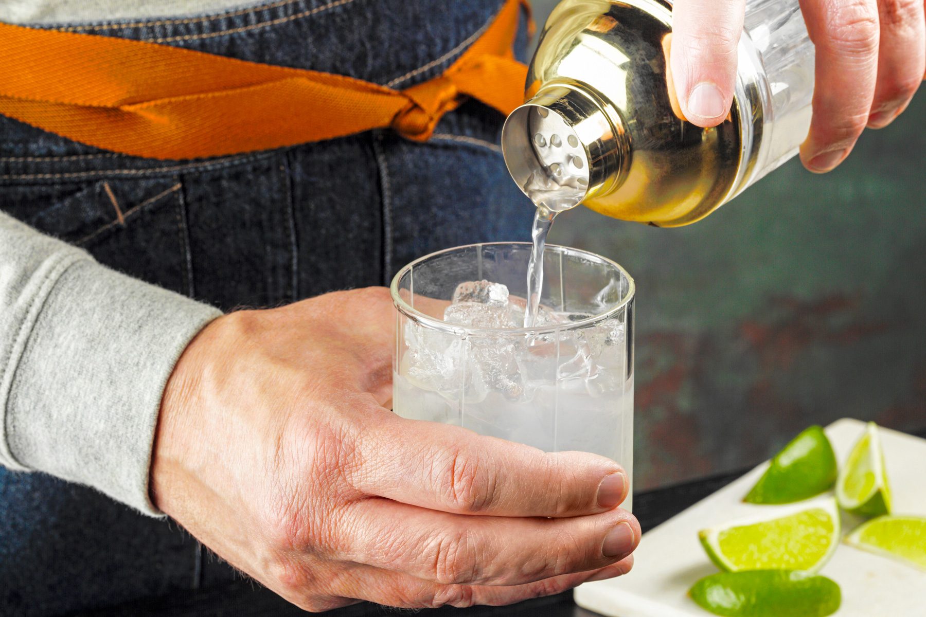 3/4 angle view shot of pouring into an ice-filled glass; lime wedges; chopping board; green and brown texture wall; black wooden base;