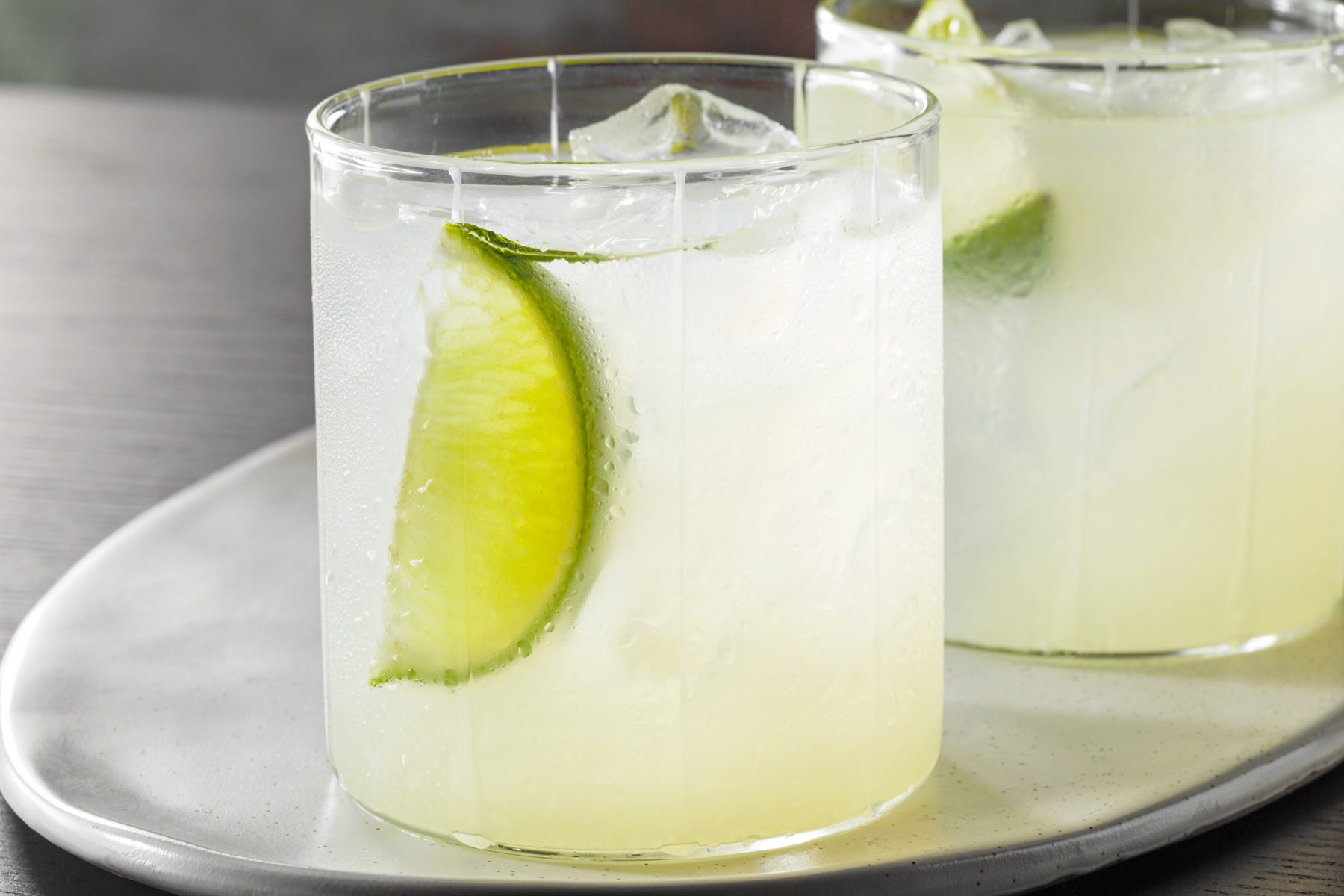 Table view shot of Gimlet; served in ice-filled glasses; garnished with lime wedges; on white tray; black wooden base