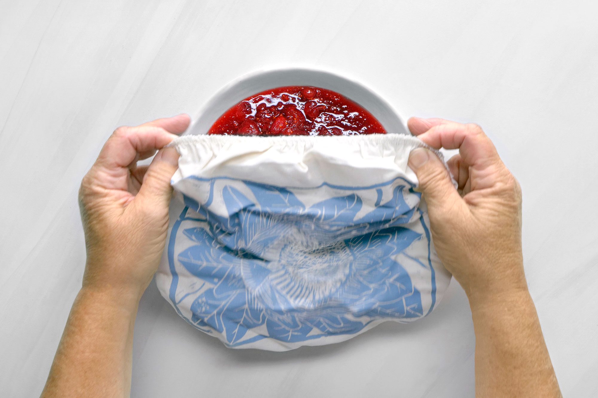 overhead shot; white background; A person is holding a white cloth over a bowl filled with cranberry sauce, The cloth has a blue and white pattern with a design of leaves, The person's hands are holding the edges of the cloth, creating a tent-like shape over the bowl, The sauce is visible through the cloth, and there are a few small bubbles on the surface;