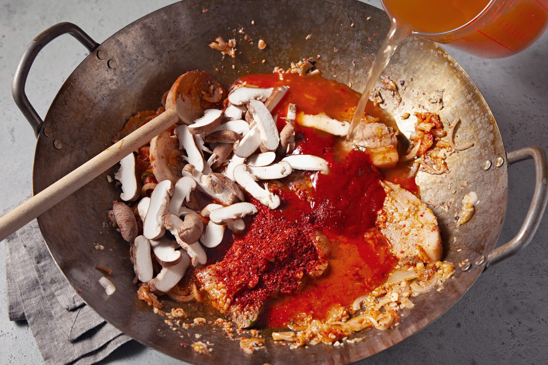 3/4th shot; light grey background; in the same wok, Stiring in vegetable broth, mushrooms, kimchi juice, gochujang, gochugaru, soy sauce and fish sauce