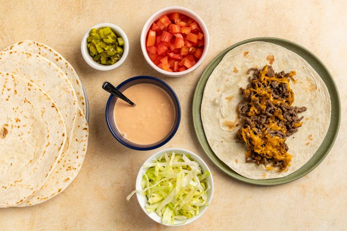 Taste of Home Cheeseburger Tacos recipe photo of the cheesy beef transferred to a tortilla with the tomato, pickles, and lettuce on side for dressing up the taco.