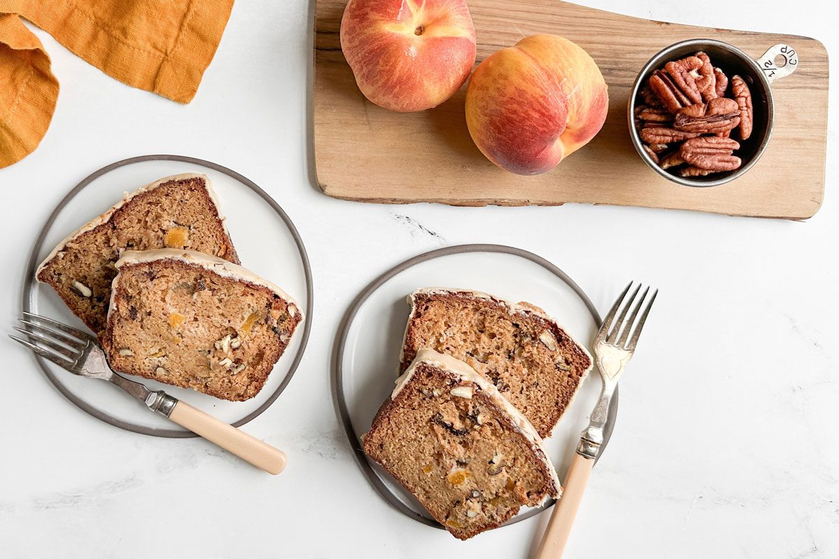 Taste of Home Peach Bread on ceramic plates next to a wooden board topped with peaches and a dish of pecans