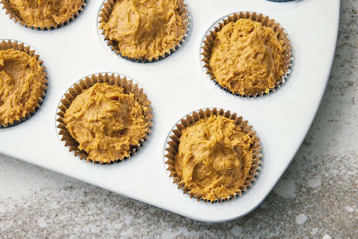 A muffin tray containing six metallic cupcake liners filled with unbaked pumpkin batter, resting on a speckled countertop. The batter has a rough, uneven texture, and the tray is positioned diagonally in the frame.