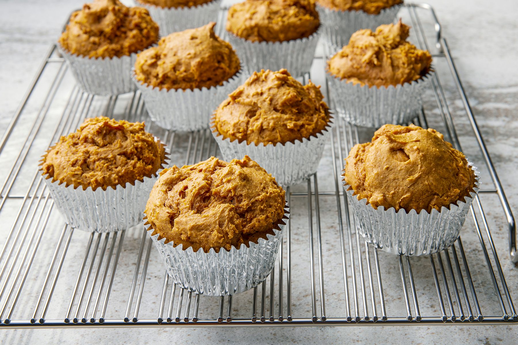 Freshly baked muffins in silver foil liners cooling on a wire rack. The muffins have a golden-brown, slightly rough texture and are set on a light-colored countertop.