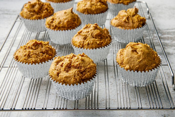 Freshly baked muffins in silver foil liners cooling on a wire rack. The muffins have a golden-brown, slightly rough texture and are set on a light-colored countertop.