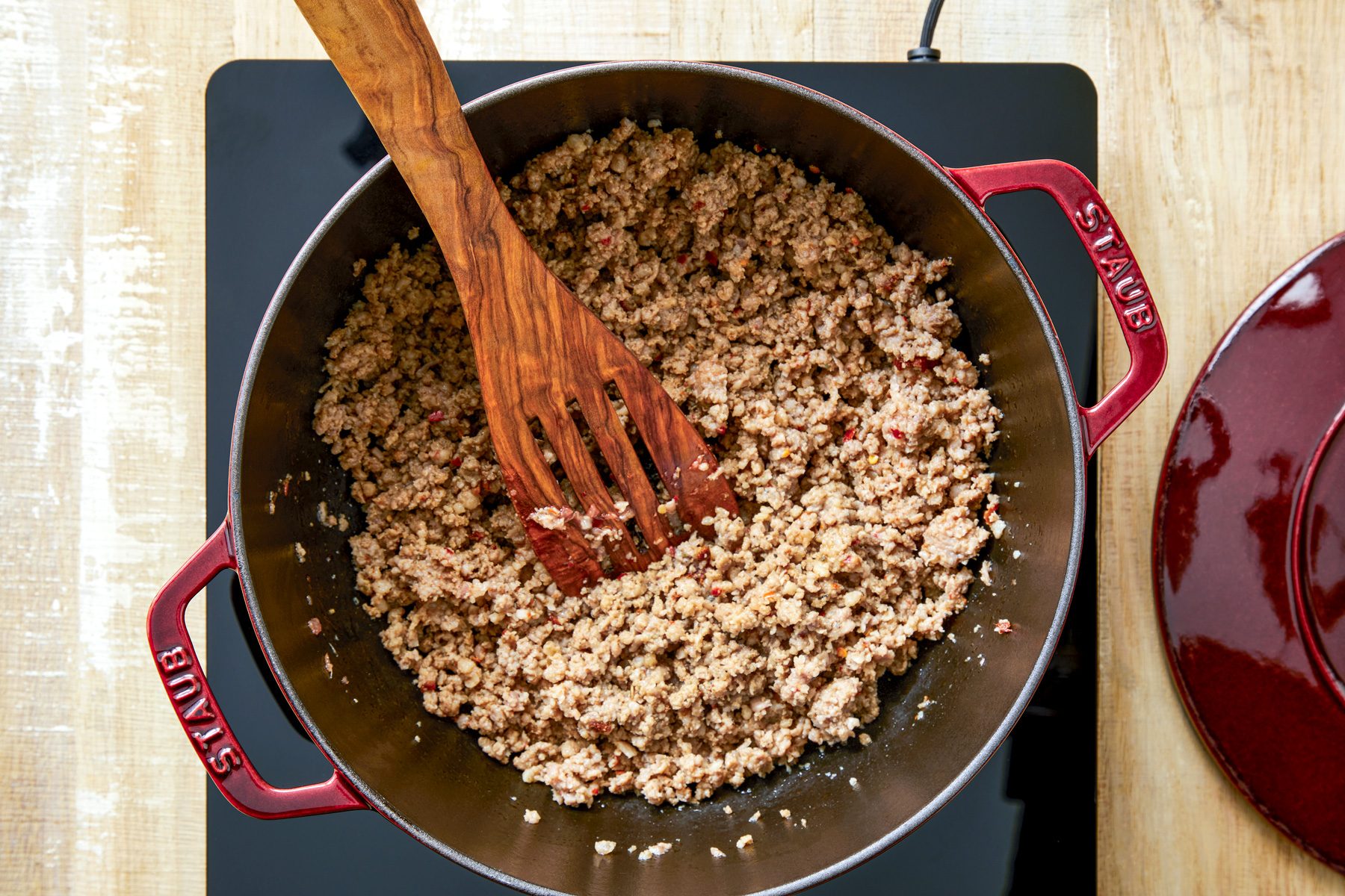 overhead shot; wooden background; In a dutch oven cooking crumbled sausage over medium heat until no longer pink; a wooden spatula rests on the pan
