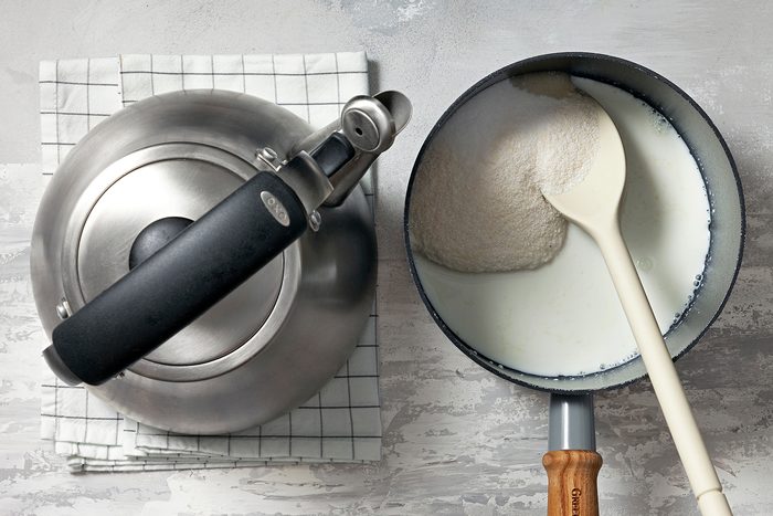 A kettle and a saucepan are placed on a checkered cloth. The saucepan contains a white liquid, likely milk, being stirred with a wooden spoon. The setup is on a light textured surface.
