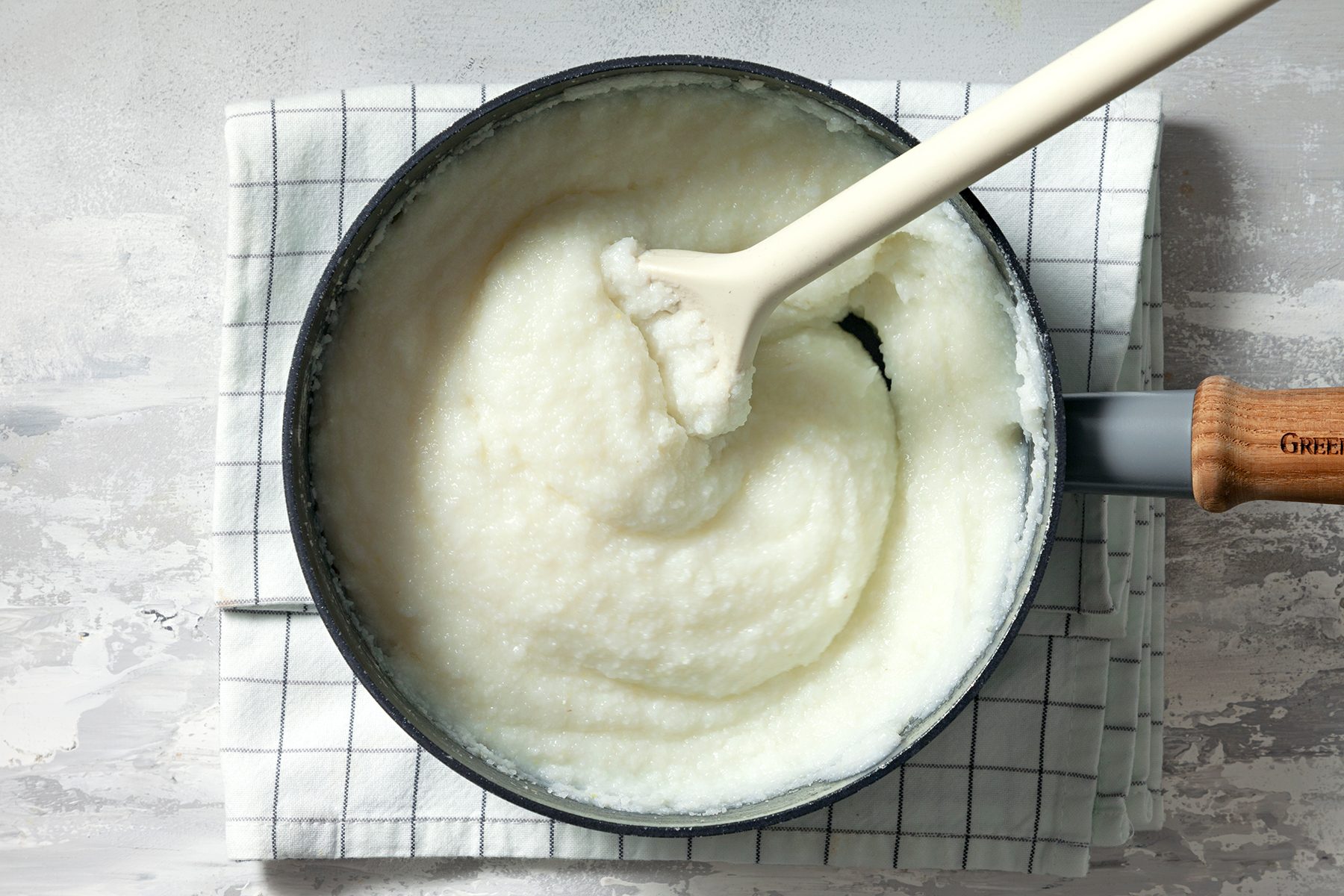 A pot of creamy, white grits is shown with a wooden-handled spoon resting inside, all placed on a checkered cloth. The smooth texture of the grits is visible, and the pot is situated on a light-colored surface.