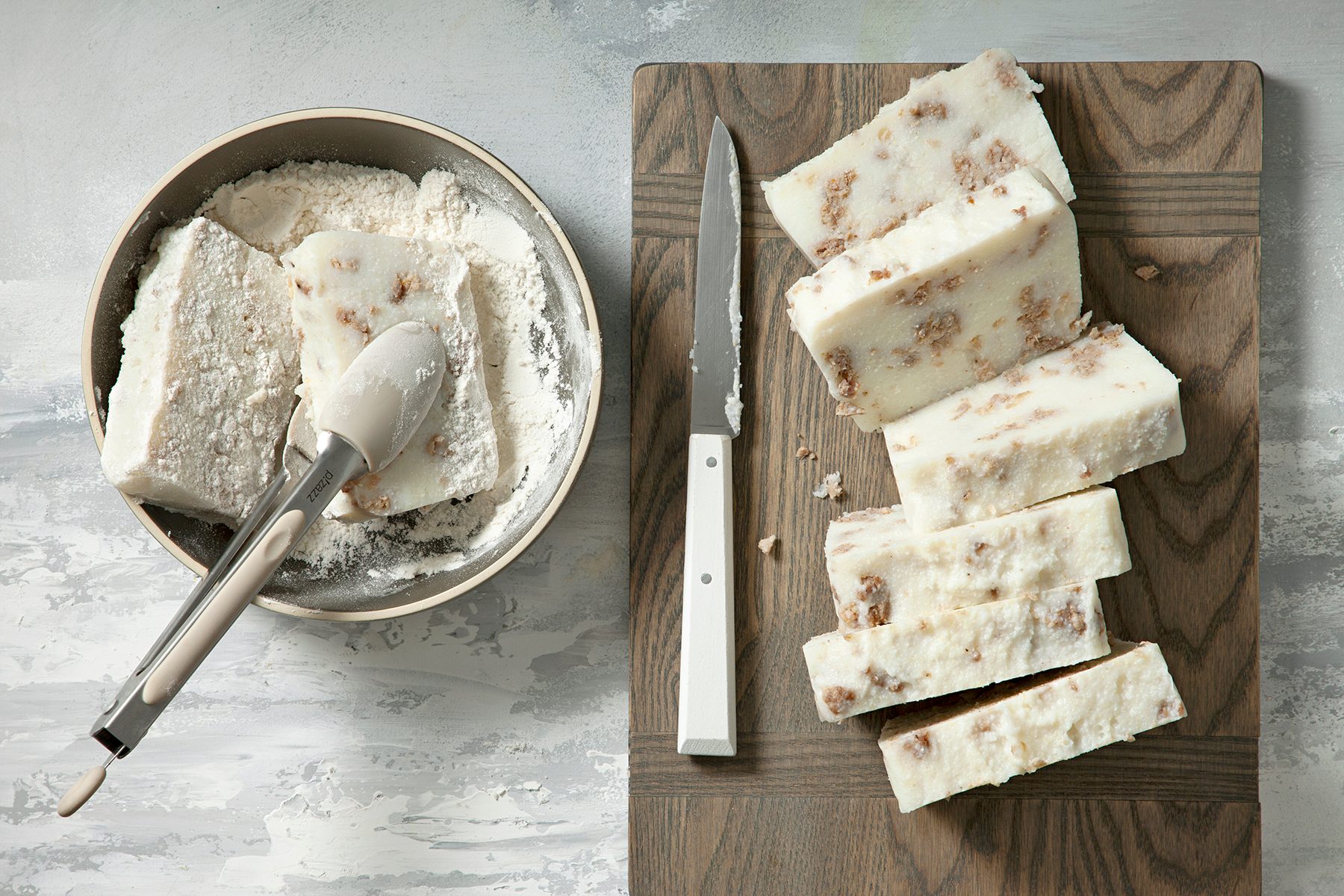 A wooden cutting board with slices of ice cream log next to a knife with a white handle. A nearby metal bowl contains more ice cream with a scoop resting inside. The ice cream has visible chunks of nuts or cookies. The surface beneath is a light-gray textured design.
