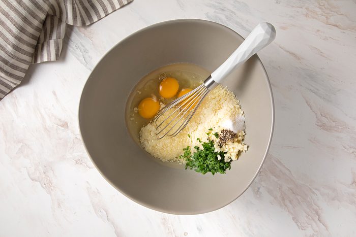 Top-down view of a mixing bowl containing three cracked eggs, grated Parmesan cheese, chopped garlic, chopped parsley, and black pepper. A whisk with a white handle rests inside the bowl. A striped cloth is partially visible in the top left corner.