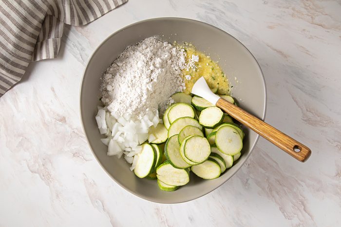 A mixing bowl on a marble countertop contains sliced zucchini, chopped onions, flour, and a whisked egg mixture. A white spatula with a wooden handle rests on the edge of the bowl. A striped kitchen towel is partially visible on the left side.