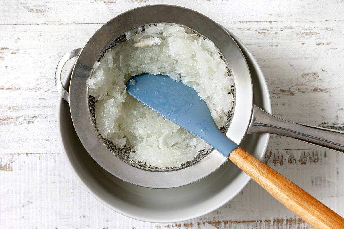 grated onion in a fine sieve over a bowl with spatula being used to press and squeeze to remove juice
