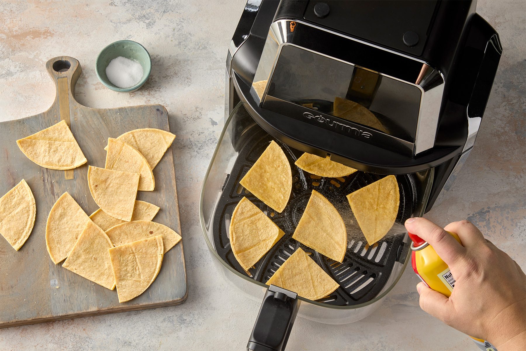 overhead shot of an air fryer with a basket full of tortilla chips, A person is spraying the chips with cooking spray, There is a wooden cutting board to the left with more tortilla chips;