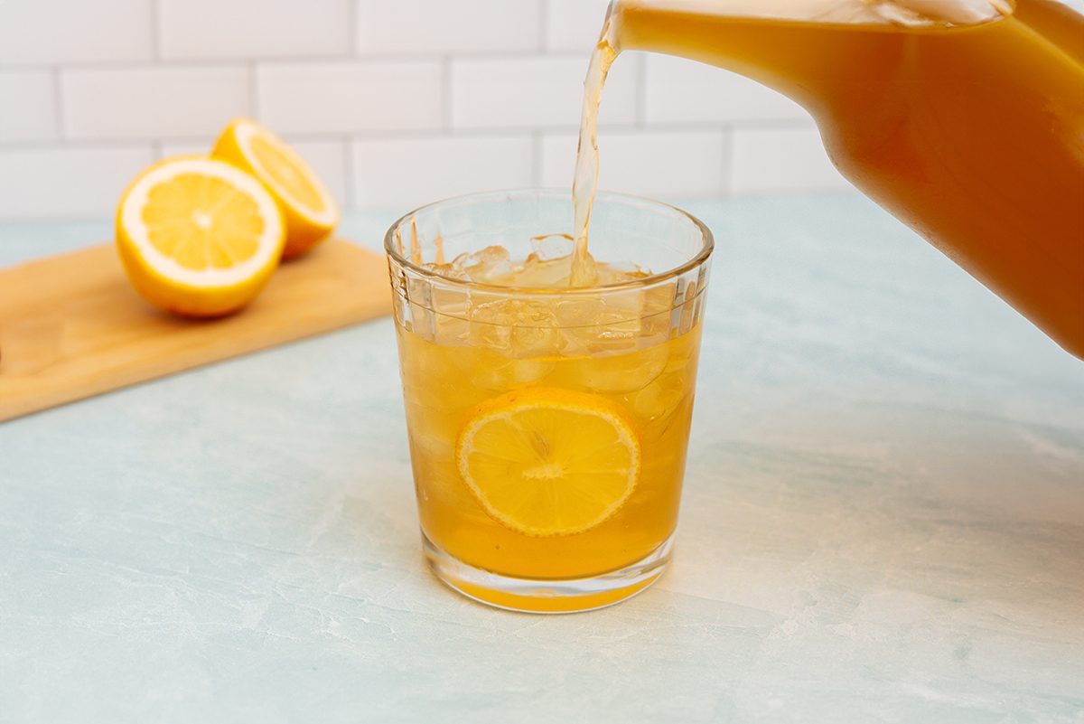 A pitcher of Arnold Palmer being poured into a glass cup with ice and a lemon in it