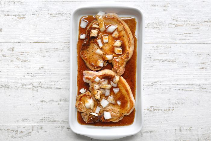 Two seasoned pork chops sit in a white rectangular dish, marinated with a sauce and topped with chopped onions. The dish is placed on a textured white wooden surface.