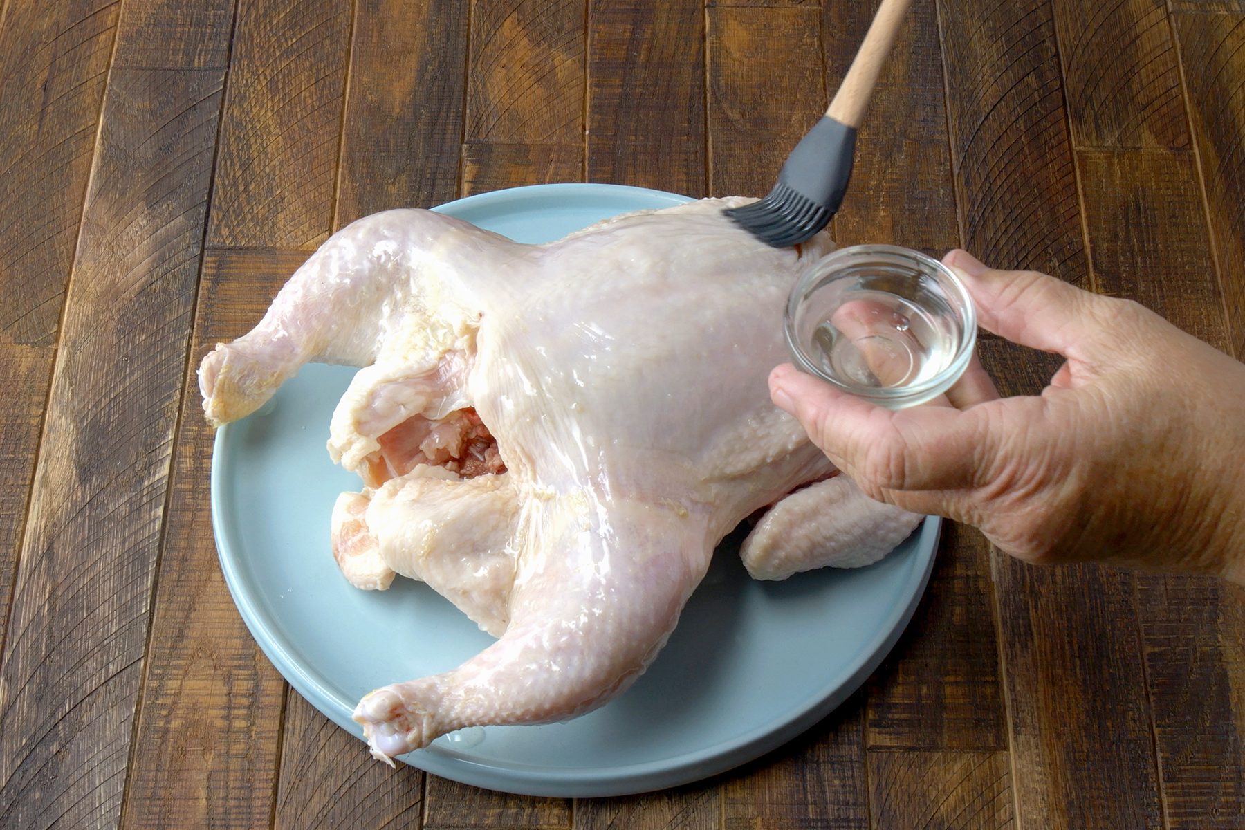 A person brushes a whole raw chicken with oil using a silicone brush. The chicken is placed on a light blue plate on a wooden surface.