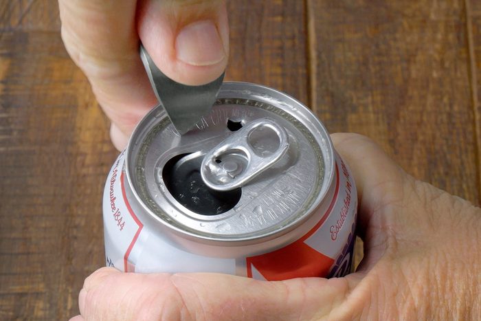 A hand is using a can opener to puncture the top of a soda can, which has a pull tab. The can is held over a wooden table surface.
