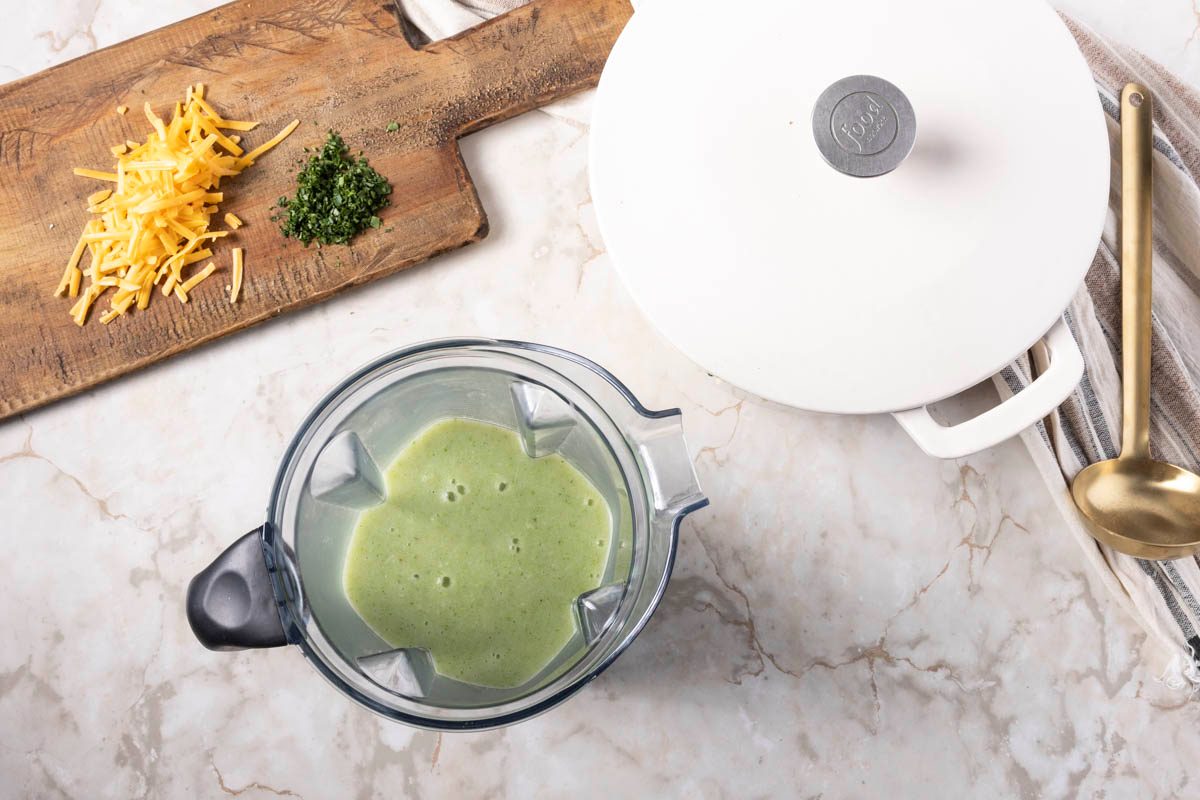 Mixing Broccoli Cheddar Potato Soup in blender next to a dutch oven on marble surface