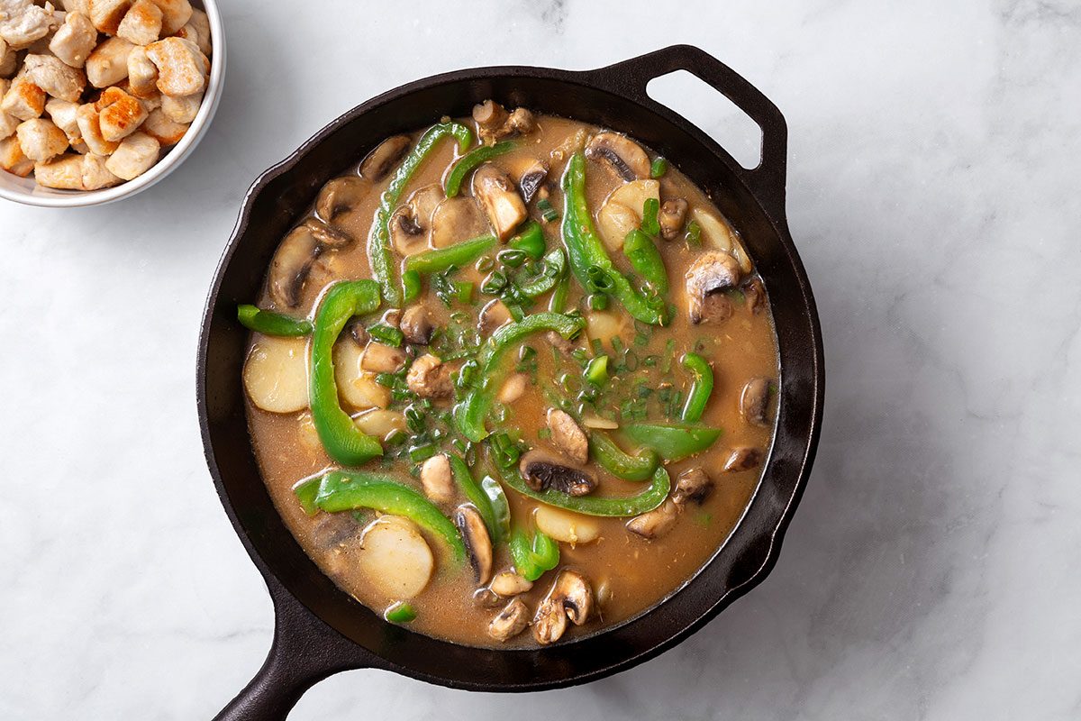 Mushrooms, green pepper, water chestnuts, and ginger root heated in a cast iron skillet with broth mixture and topped with green onion for step two of Cashew Chicken recipe for Taste of Home