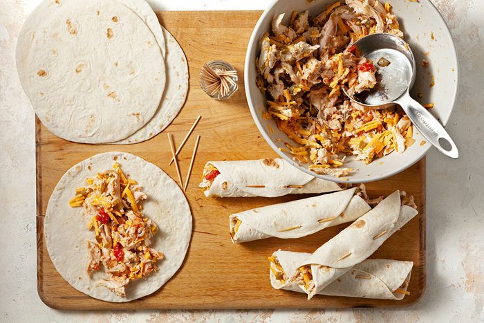 A wooden board displays tortillas being filled with a chicken, cheese, and pepper mixture. Some tortillas are rolled and secured with toothpicks, while others and the filling in a bowl. A metal scoop rests in the bowl.