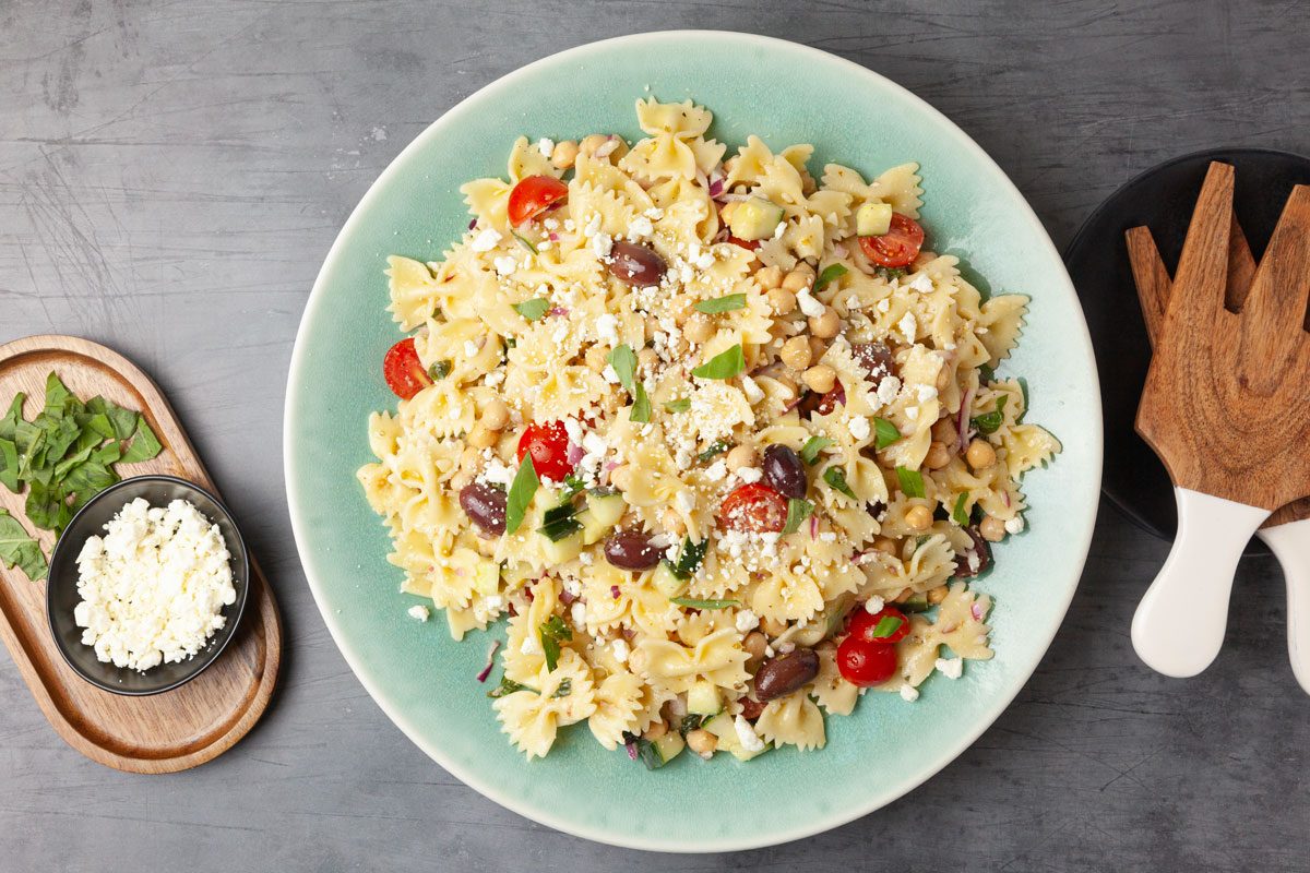 Full yield of Taste of Home Chickpea Pasta Salad in a light blue serving bowl, wooden salad servers, feta and basil