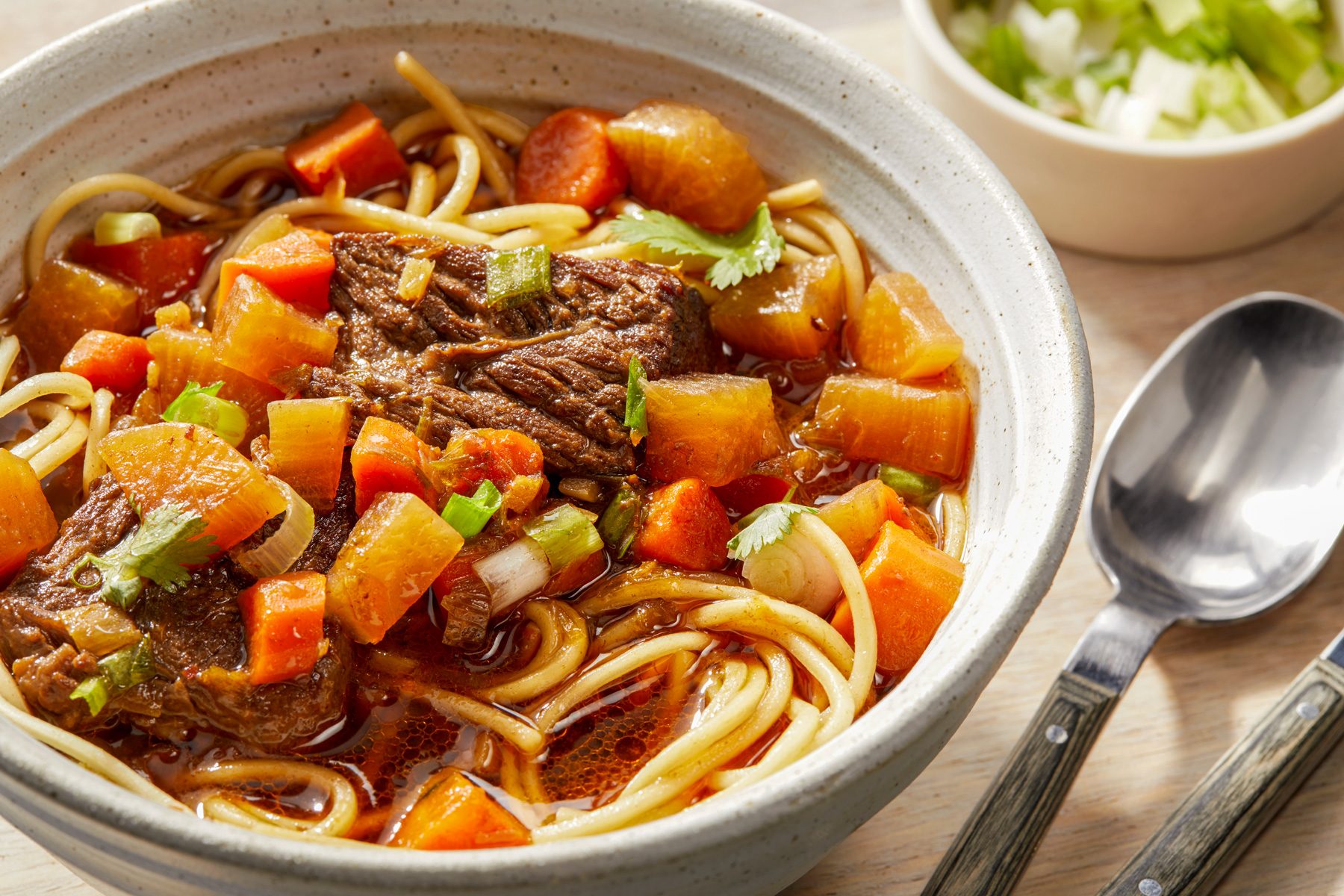 Horizontal Push shot of A bowl of hearty beef noodle soup with carrots, potatoes, and green onions, A spoon and a fork with wooden handles are beside the bowl, The bowl is sitting on a wooden surface.