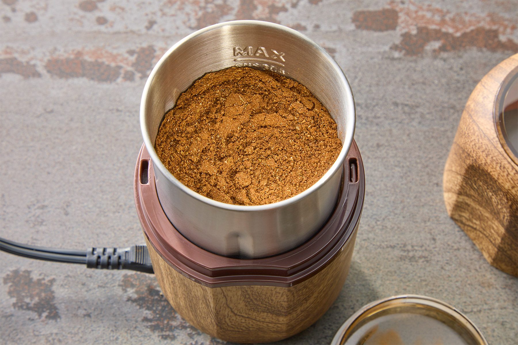 A metal cup filled with ground coffee sits inside a wooden base of a coffee grinder. The grinder has a power cord attached. The surface underneath is textured and gray.