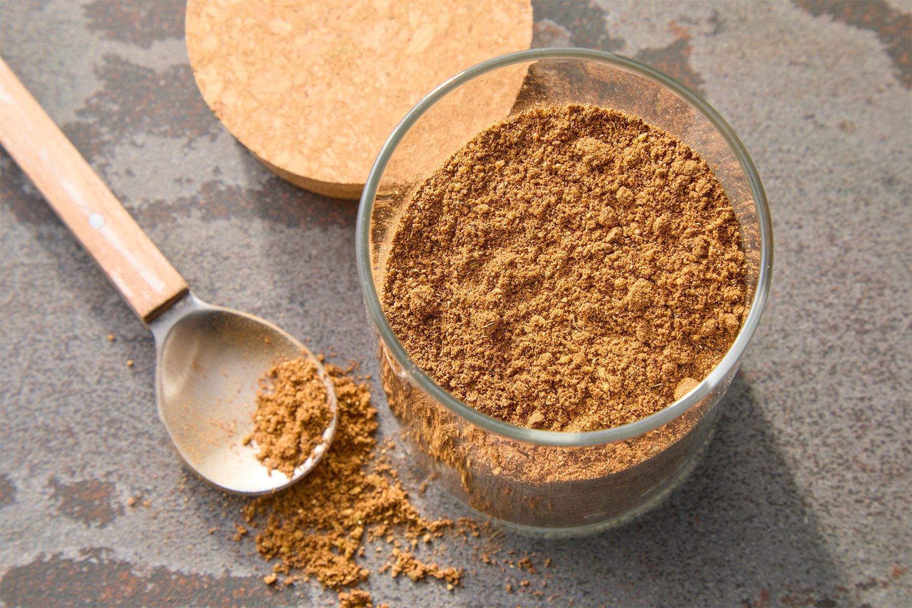 A glass jar filled with ground cinnamon sits next to a spoon with a small amount of the spice spilled on a gray surface. A wooden lid rests nearby.
