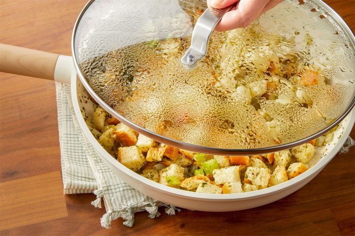 3/4th shot of a white pan filled with a mixture of diced bread, chopped celery, and chopped onions, The bread cubes are a light brown color, and the vegetables are a mix of green and white, A hand is lifting the lid off the pan, revealing a cloud of steam;