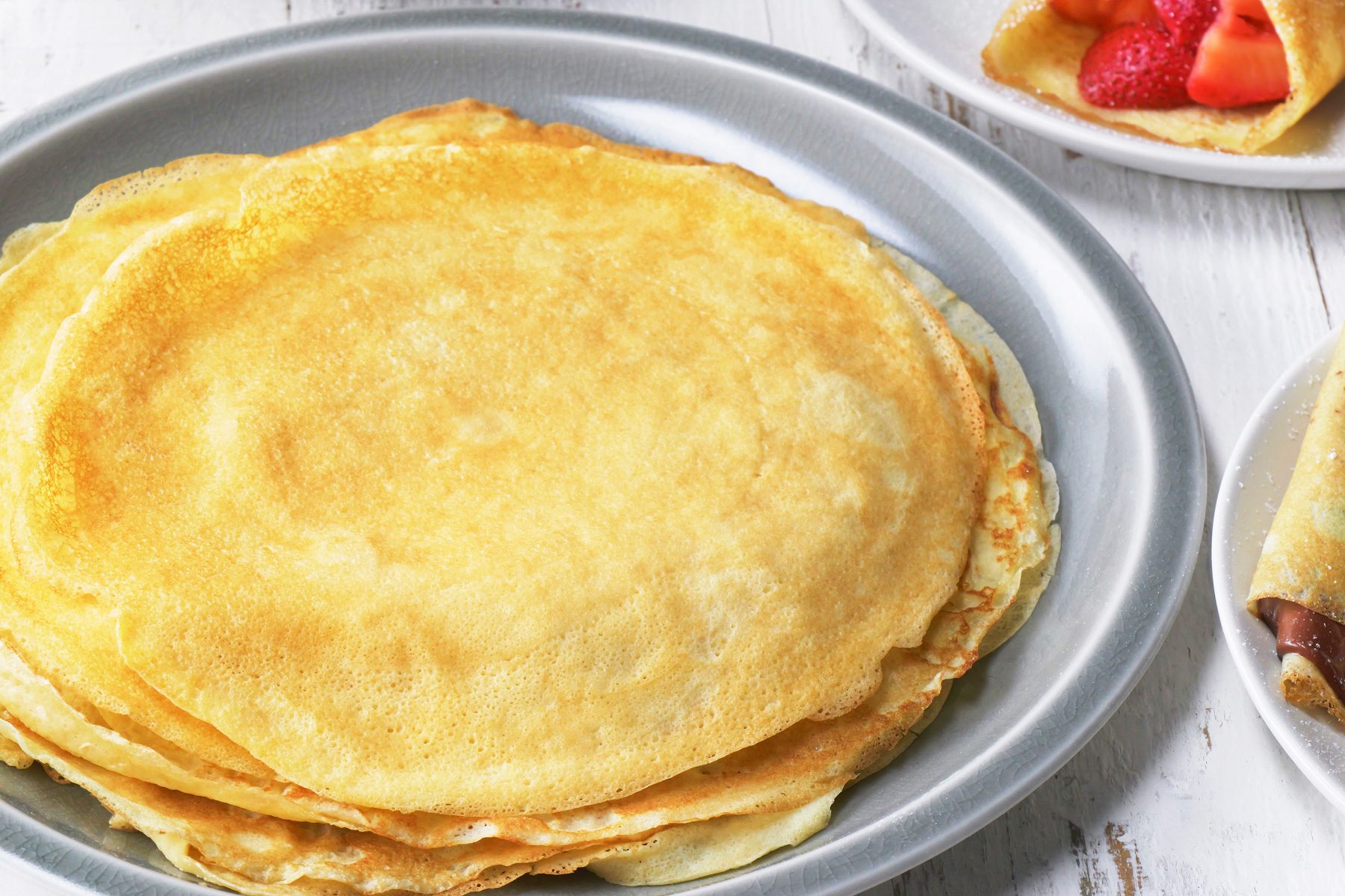 3/4th shot; white wooden background; a stack of crepes on a plate, The crepes are thin and golden-brown with slightly crisp edges, In the background, there is another plate with a single crepe folded and filled with strawberries