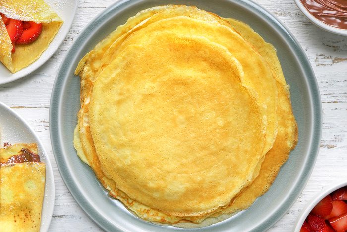 overhead shot; white wooden background; a stack of crepes on a plate, The crepes are thin and golden-brown with slightly crisp edges, In the background, there is another plate with a single crepe folded and filled with strawberries