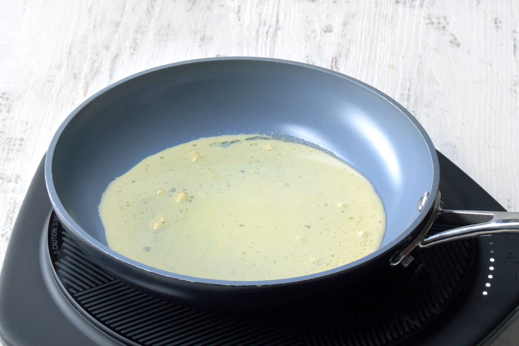 3/4th shot; white wooden background;  a blue frying pan on a stove, Inside the pan, there’s a liquid mixture, possibly batter for pancakes or crepes, with small lumps