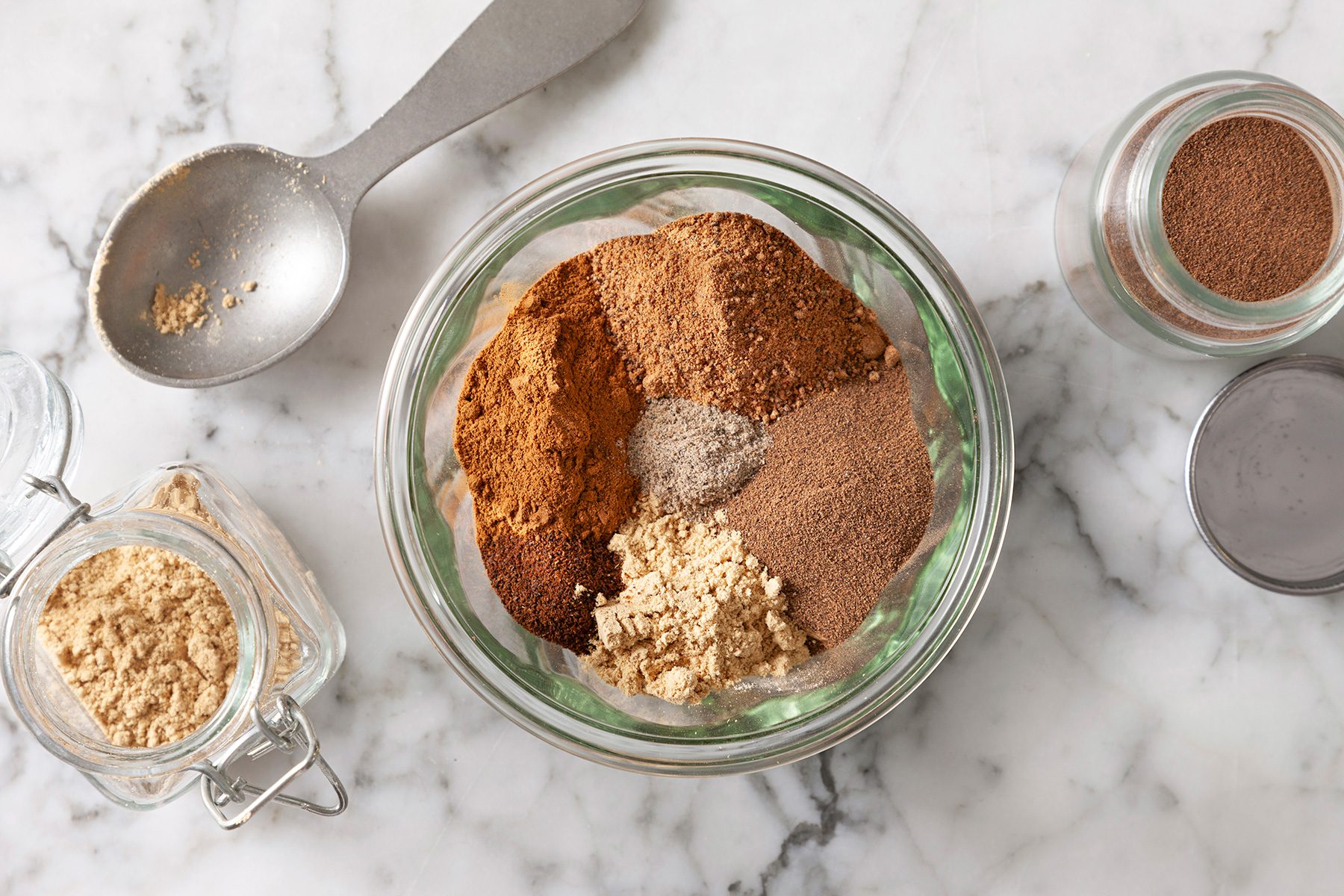 A glass bowl filled with various ground spices, including cinnamon and ginger, on a marble surface. Nearby are small jars of additional spices and a metal spoon. The scene suggests a homemade spice blend preparation.