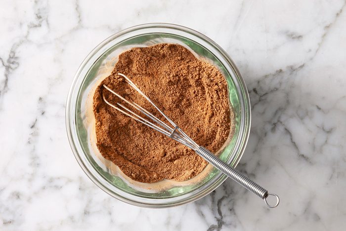 A clear glass bowl filled with brown spice mix, placed on a marble surface. A metal whisk rests inside the bowl.
