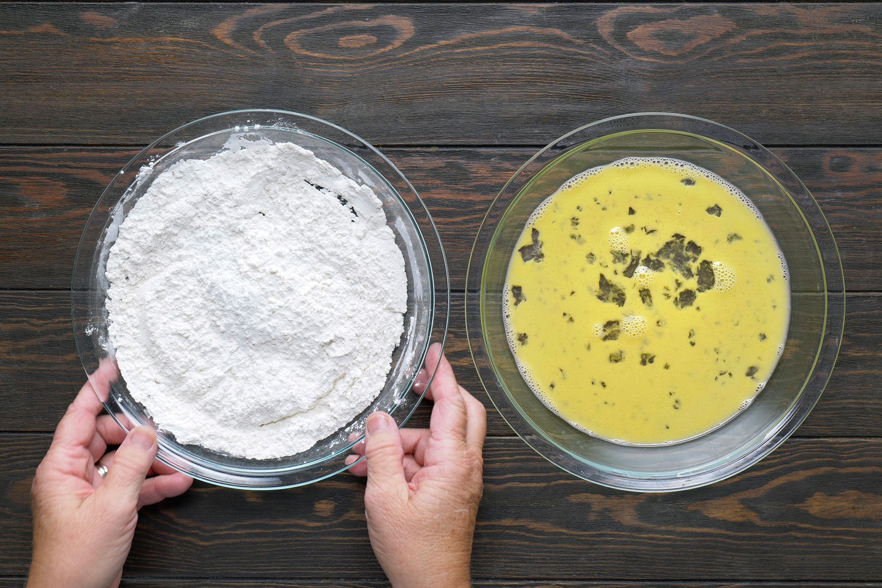 Two bowls on a wooden surface: the left bowl contains flour, and the right bowl has a yellow egg mixture with herbs. Hands are holding the flour bowl.