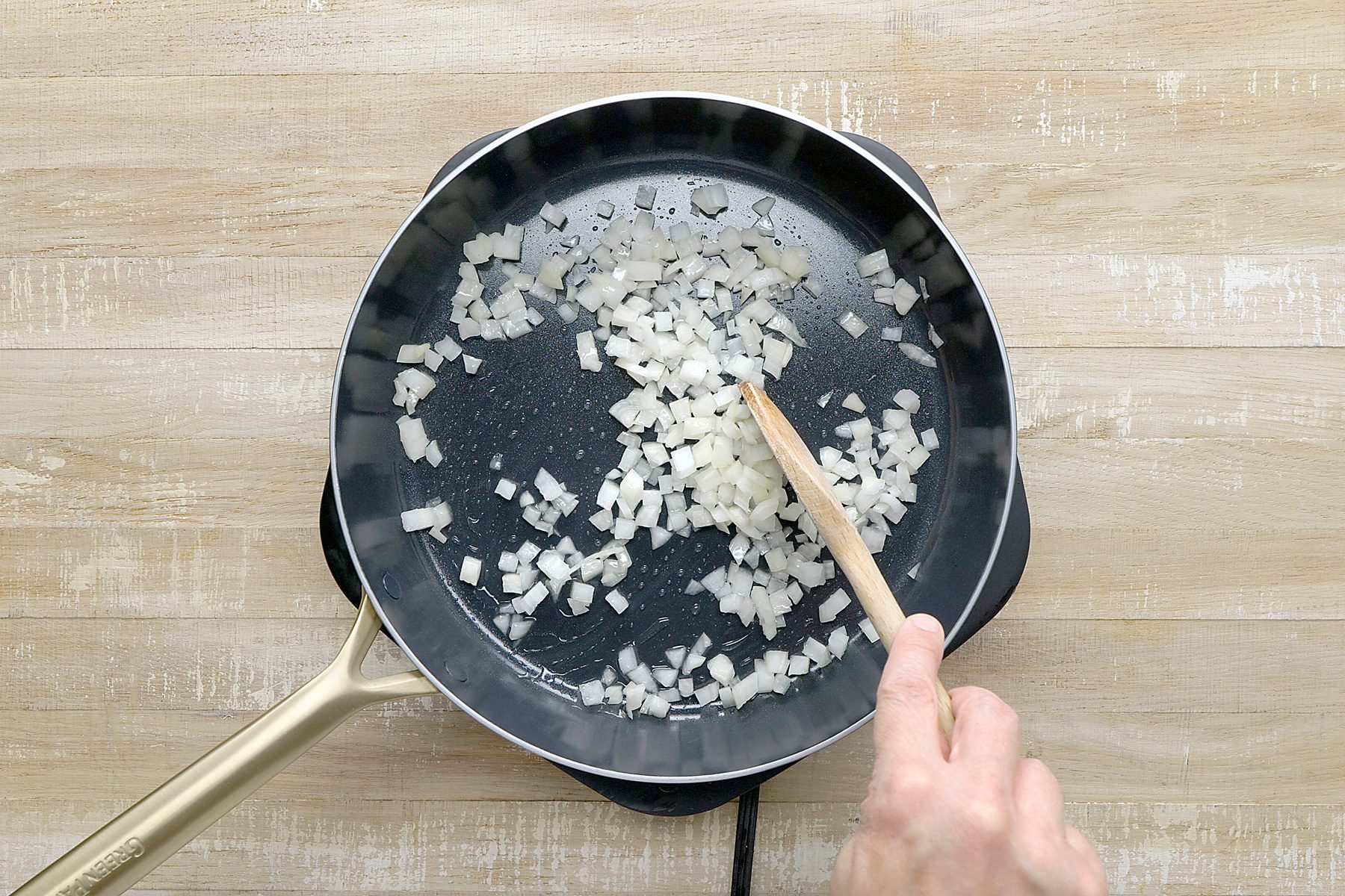 overhead shot of a black skillet on an electric induction cooktop, The skillet contains chopped onions that are being sautéed in oil or butter; The onions are translucent and are sizzling slightly, A wooden spatula is being used to stir the onions;