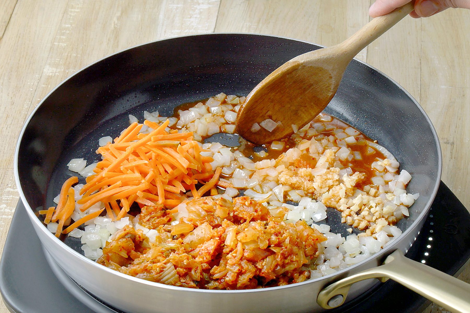 3/4th shot of a black skillet contains a mixture of chopped vegetables and kimchi, The vegetables include shredded carrots, chopped onions, and minced garlic, The kimchi is a vibrant red color, A wooden spatula is being used to stir the ingredients together;
