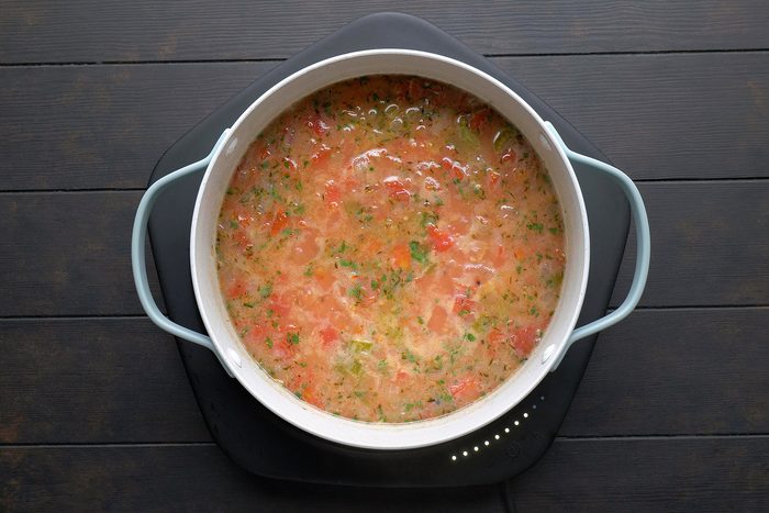 A pot of soup on a black cooking surface, containing a mix of chopped vegetables such as tomatoes, herbs, and other ingredients, creating a colorful and hearty dish. The pot has light-colored handles.