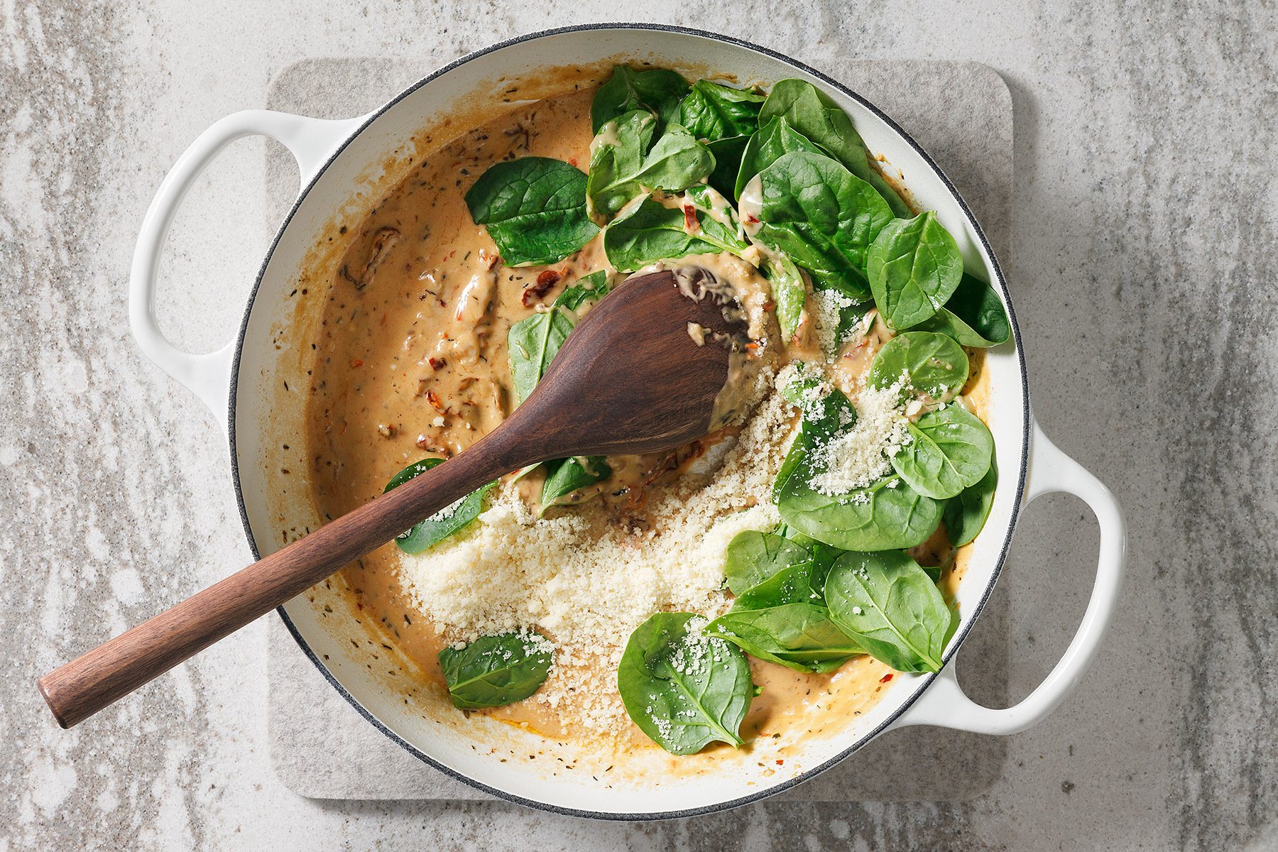 overhead shot of a white pot with a wooden spoon lying across the top of the pot, The pot contains a creamy sauce with spinach and grated cheese on top, The pot is sitting on a light grey countertop;