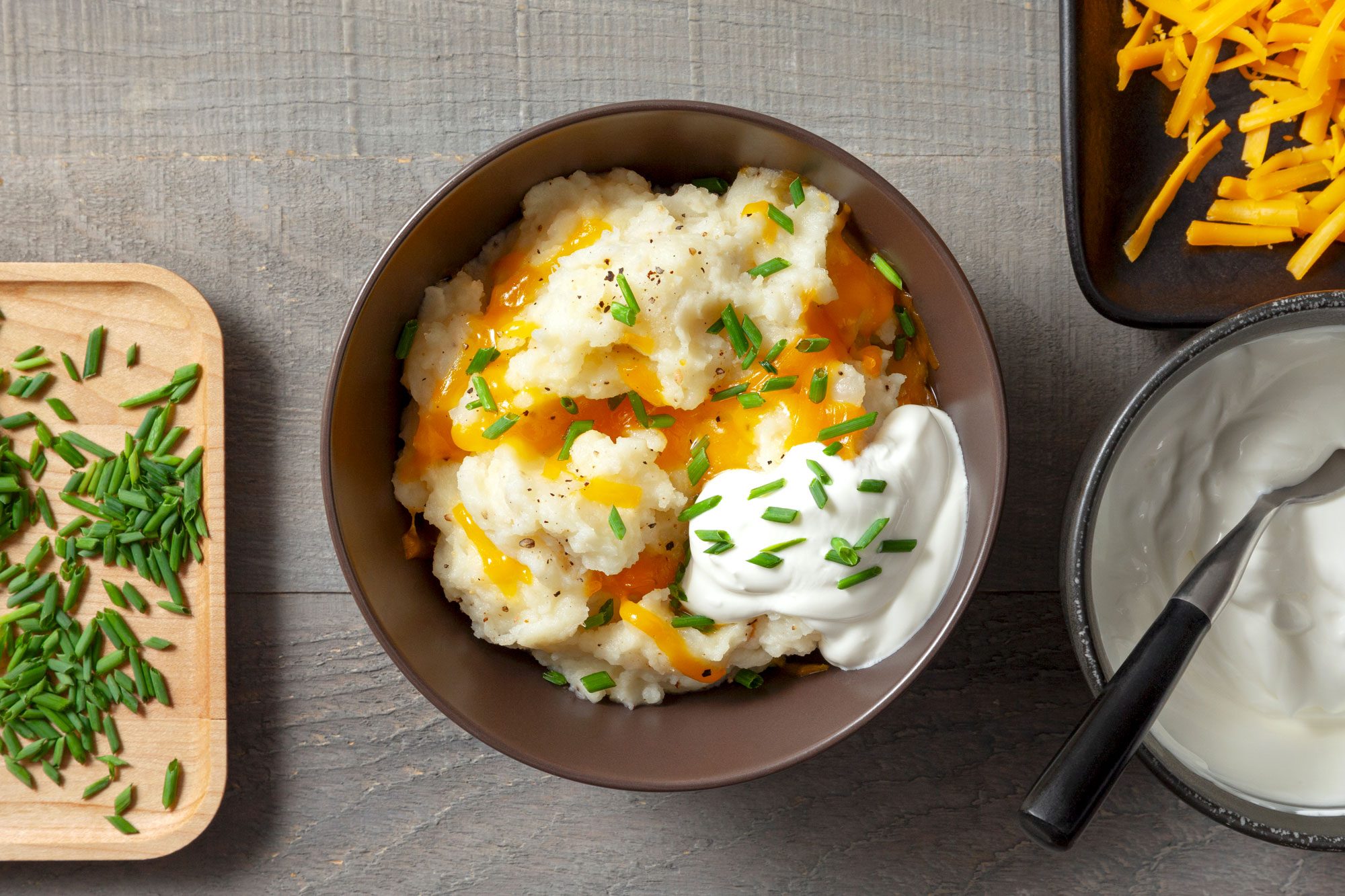 Overhead shot of Mashed Potato Bar; serve in second bowl; topping with cheese; sour cream and chopped fresh shives; spoon; wooden surface;