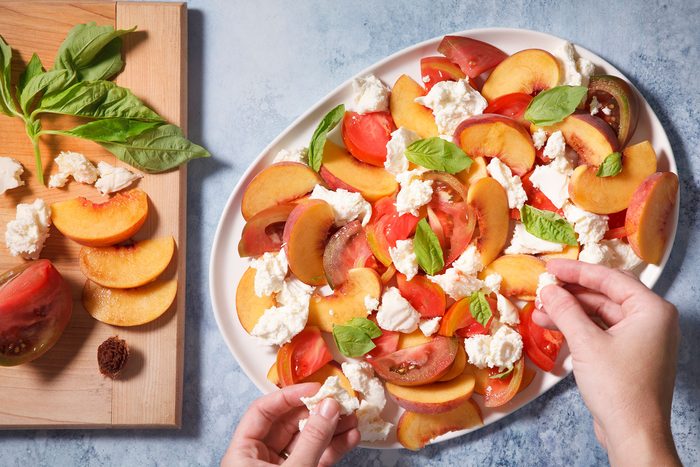 A platter with sliced peaches, tomatoes, and dollops of mozzarella topped with fresh basil leaves. A person is adding cheese to the salad, and more ingredients are on a wooden board beside the dish.