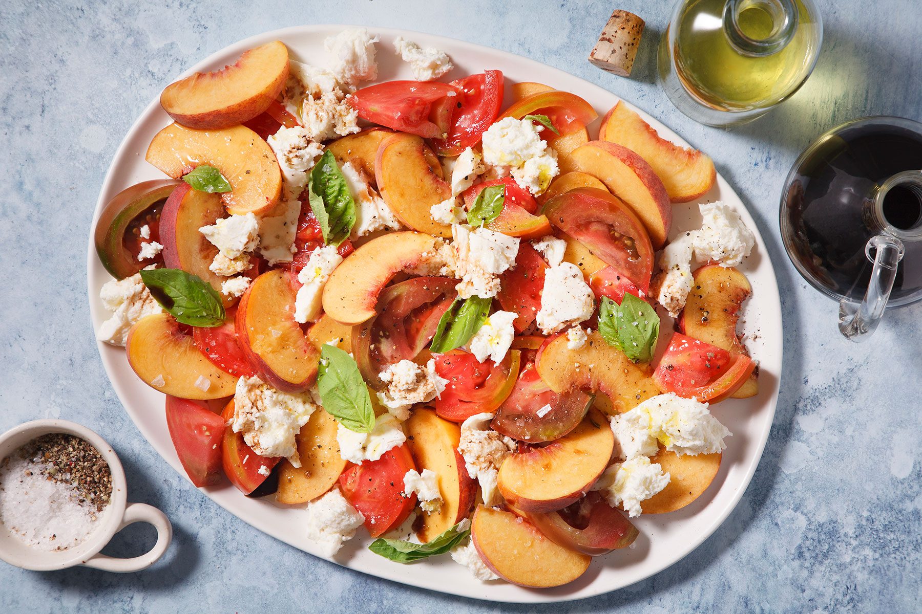 A colorful platter of sliced peaches, tomatoes, and chunks of mozzarella cheese is garnished with fresh basil leaves. Nearby are a small bowl of salt and pepper, a bottle of olive oil, and a cork with a shadow on the blue surface.