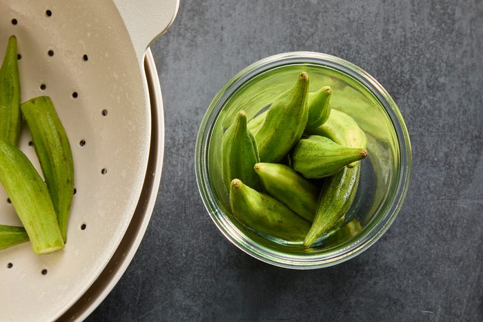 A glass jar filled with fresh green okra on a dark gray surface, next to a white colander containing more okra. The scene suggests preparation for cooking or preserving the vegetables.