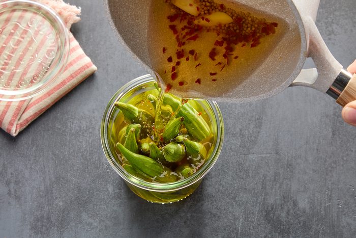A hand pours a spiced brine from a pot into a jar filled with fresh okra. A red-striped cloth and a lid are nearby on a dark surface. The brine contains visible crushed red pepper and mustard seeds.