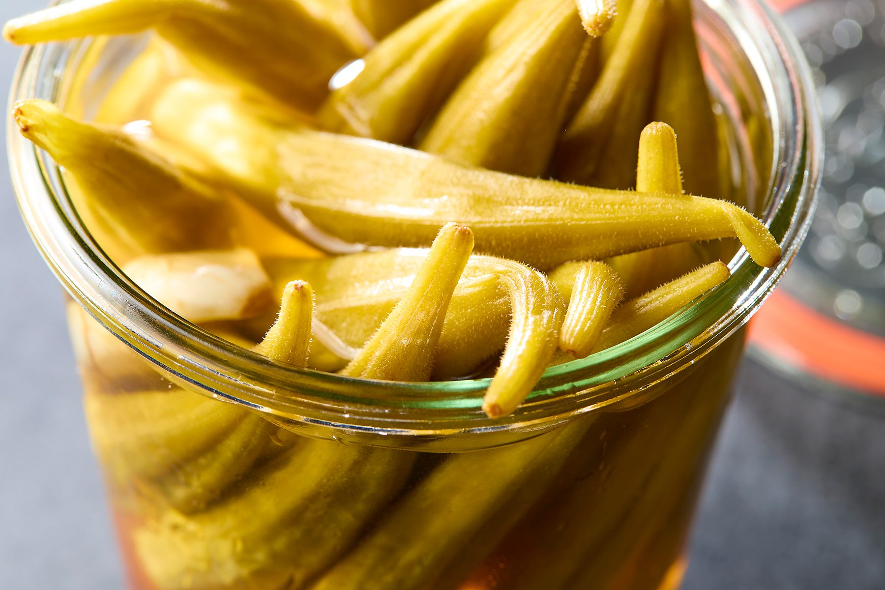 A close-up of a jar filled with pickled okra, with the green vegetables submerged in a light yellowish brine. The jar is placed on a dark surface, capturing the texture and details of the okra and the glass.