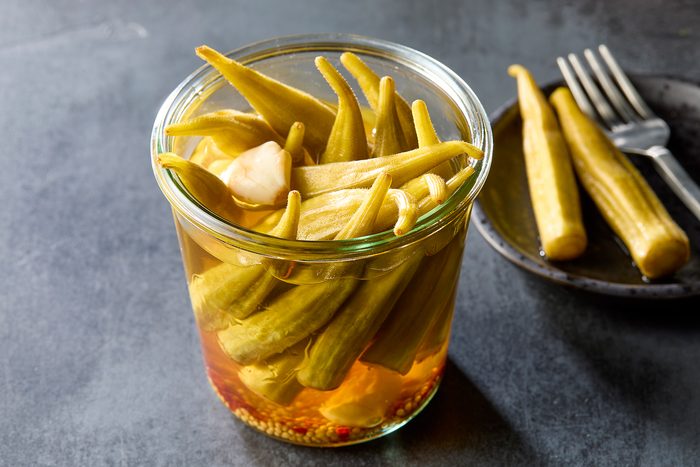 A jar of pickled okra with cloves of garlic and spices sits on a dark surface. Nearby, a small plate holds a few pieces of okra with a fork. The okra is submerged in a golden liquid, showcasing its bright green color.