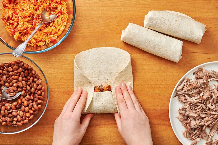 Folding a burrito with Spanish rice and beans in bowls and shredded meat nearby