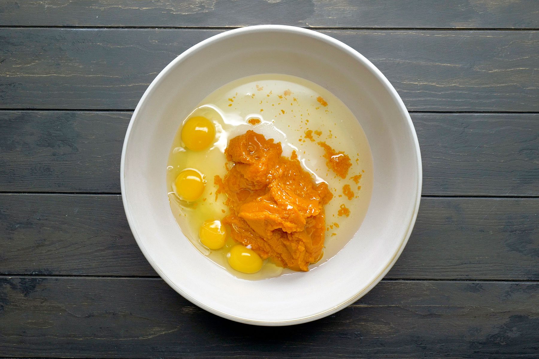 A white bowl on a dark wooden surface contains a mixture of raw eggs, egg yolks, and pumpkin puree.