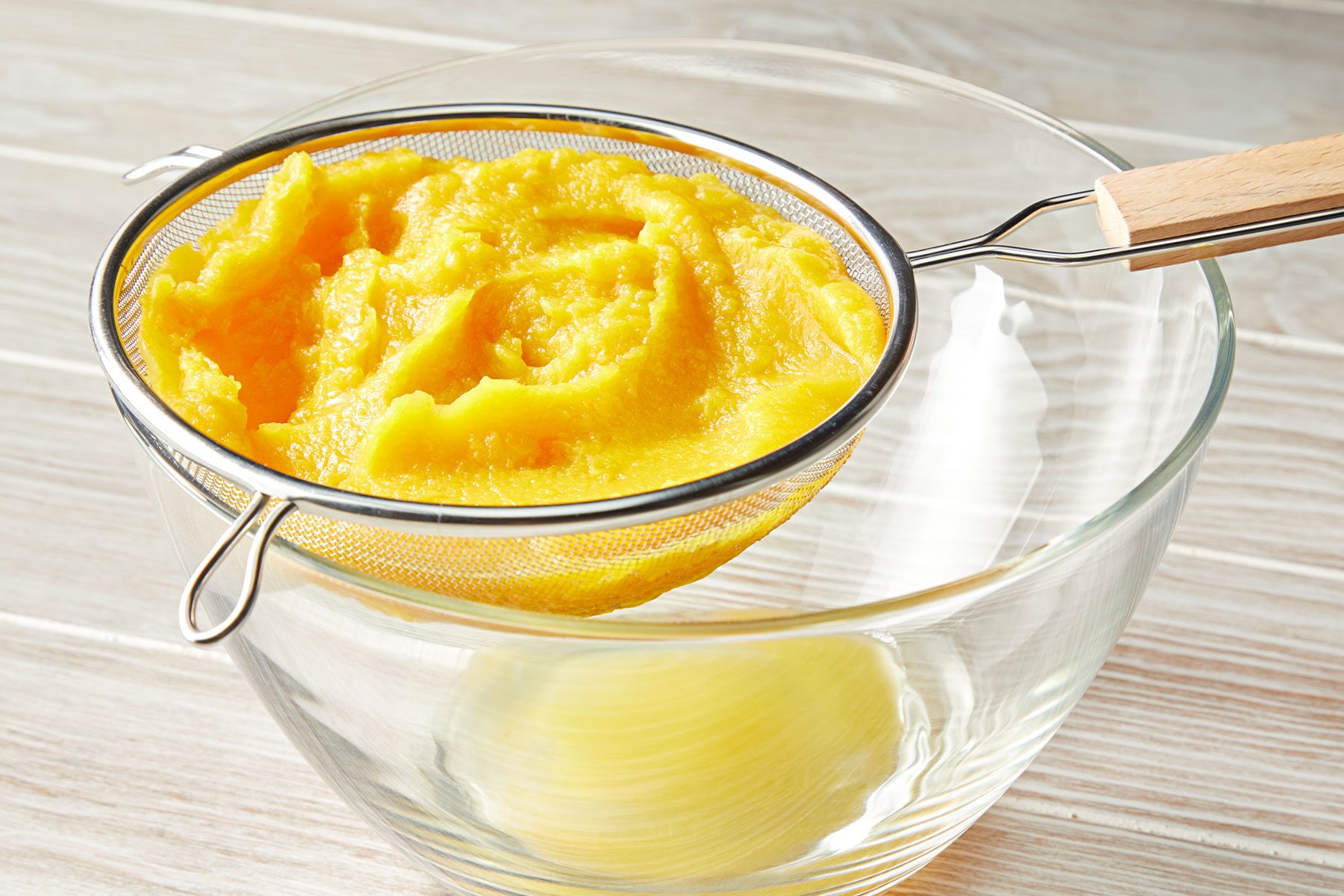 Puréed pumpkin in a mesh strainer over a clear glass bowl on a wooden surface.