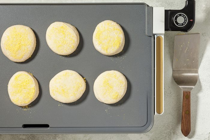 overhead shot of a griddle with six round dough circles arranged in a grid pattern, The dough circles are coated in cornmeal, A spatula is placed on the side of the griddle;